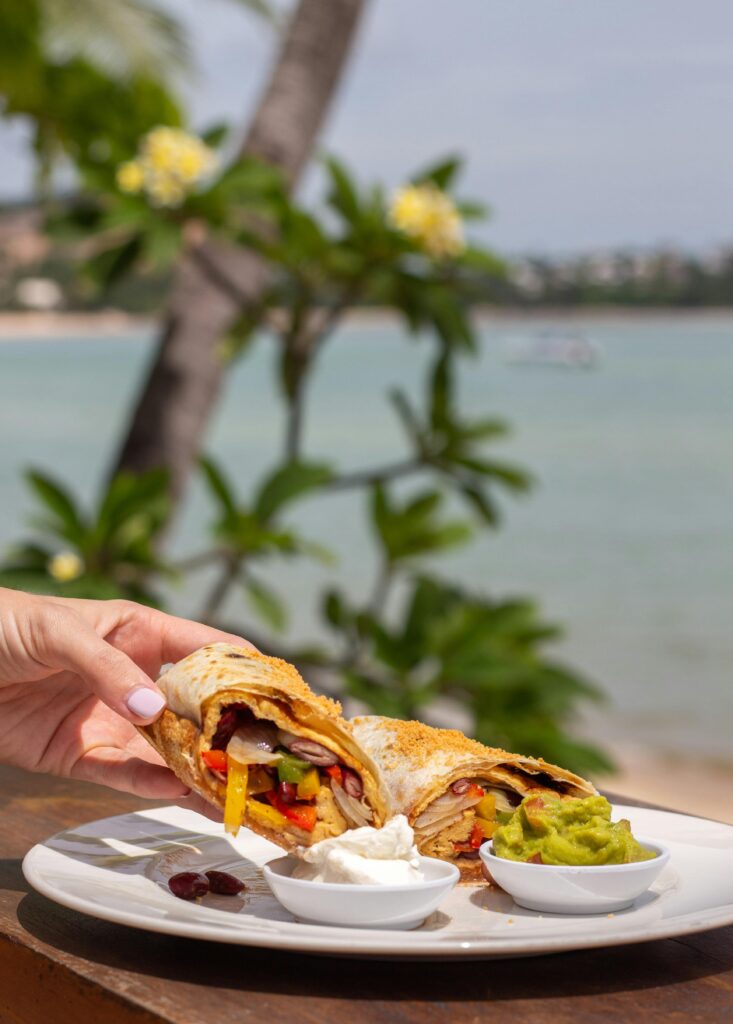 A vibrant burrito with fillings and guacamole by a tropical beach setting.